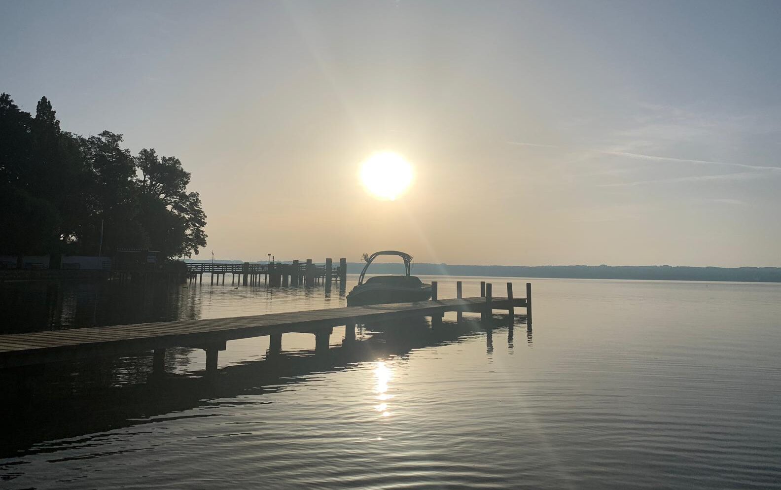 Blick vom Ufer auf See mit Steg und dort liegendem Segelboot bei Sonnenaufgang